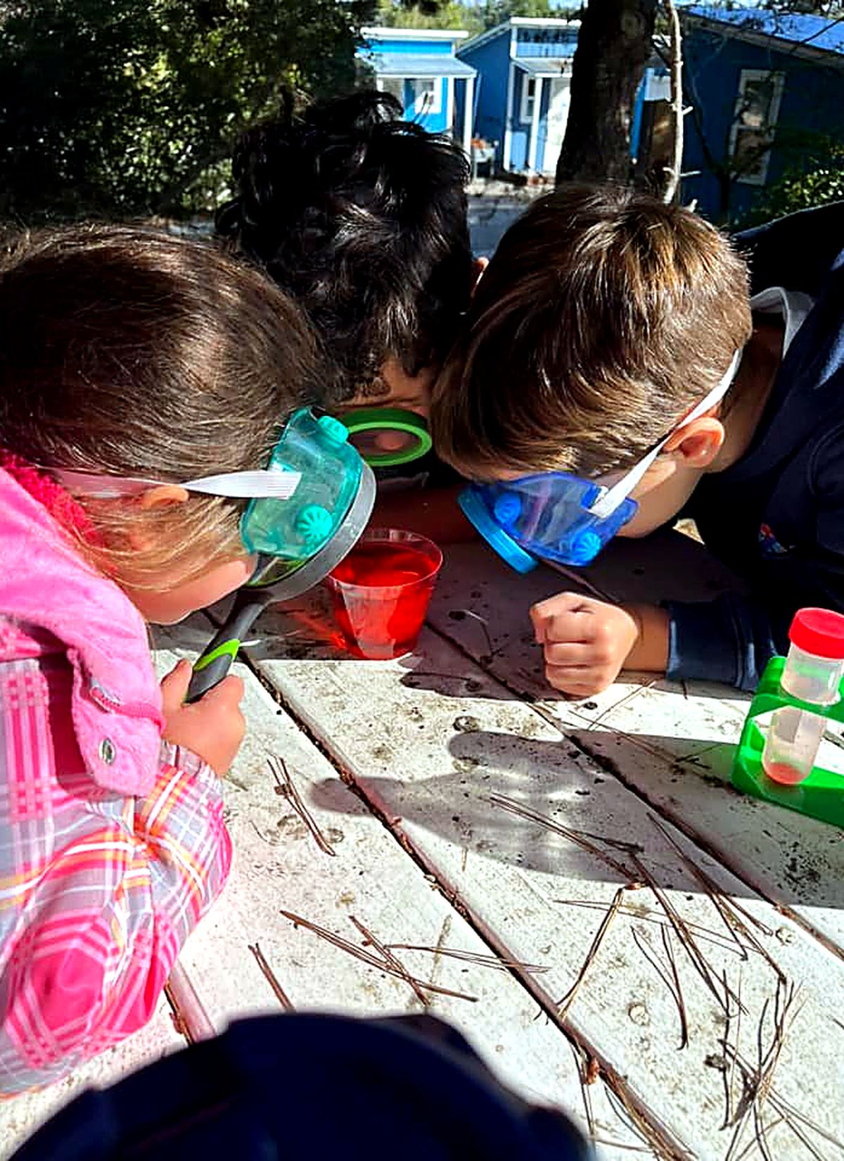Three children in safety goggles leaning over a science experiment at an outdoor table at Big Tree Little Preschool in Vista, California